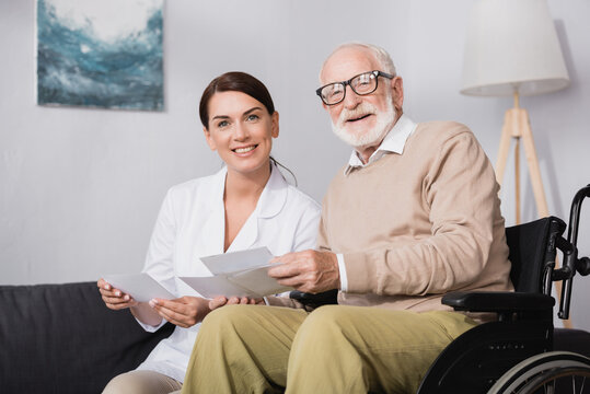 smiling social worker and aged man looking at camera while holding photos