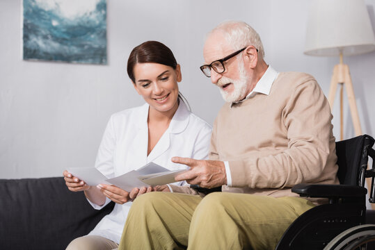 cheerful elderly man and smiling social worker looking at photos at home