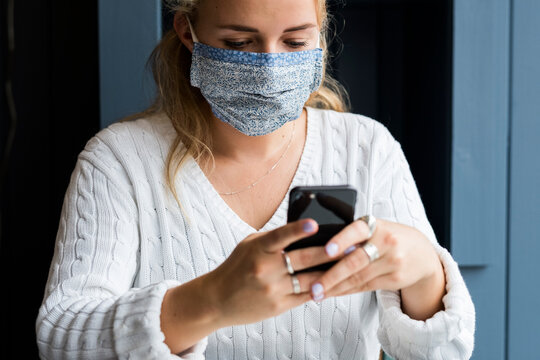Young blond woman wearing face mask sitting alone in a cafe, using mobile phone, working remotely.