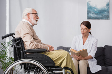 geriatric nurse reading book to aged handicapped man on blurred background