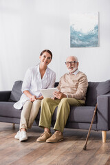 cheerful aged man holding digital tablet near smiling social worker while sitting o sofa at home