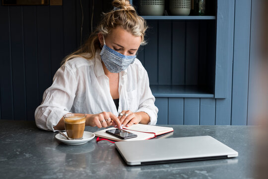 Woman wearing face mask sitting alone at a cafe table with a note book and calculator, working remotely.