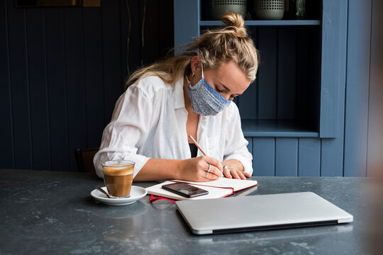 Woman wearing face mask sitting alone at a cafe table writing in a notebook working remotely.