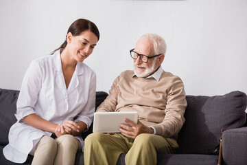 smiling aged man using digital tablet near cheerful geriatric nurse at home