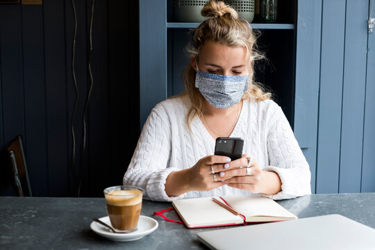 Woman wearing face mask sitting alone at a cafe table with a laptop computer, using mobile phone