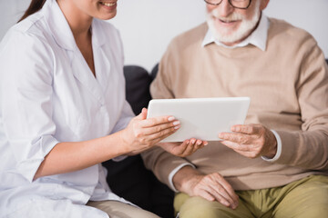 cropped view of geriatric nurse showing digital tablet to aged man on blurred background