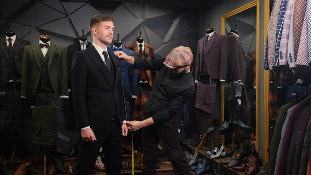 A bearded tailor takes measures of a handsome young man for a new quality handmade suit in an atelier. Creative adult stylist assists the client in choosing a costume 4k