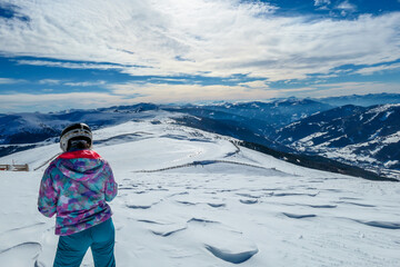 Fototapeta na wymiar A woman standing on the snow on top of Katschberg in Austria. She is happy and joyful. Panoramic view on the surrounding mountains. Winter wonderland. Bright and sunny winter day. Love and happiness