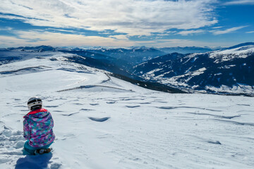 A woman sitting on the snow on top of Katschberg in Austria. She is happy and joyful. Panoramic view on the surrounding mountains. Winter wonderland. Bright and sunny winter day. Love and happiness