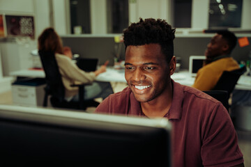 African male business man smiling while typing on desktop sitting in office with colleagues 