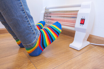 A woman wearing bright rainbow-colored socks warms her frozen feet near a home electric heater....