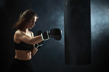 sporty brunette woman in boxing gloves and sportswear punching bag on dark background with smoke.