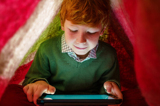 Horizontal view of little caucasian kid hidden under the bed blanket to use internet. Technology parental control for children at home. Technological lifestyle with family indoors.