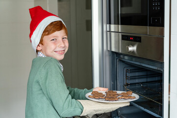 Horizontal view of red hair caucasian little boy baking fresh cookies in the oven at home with Christmas hat. Christmas traditions family recipe. Seasonal Christmas lifestyle at home with children.