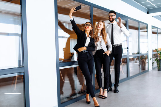 Group of attractive business people standing and chatting together holding coffee cups and taking selfie on mobile phone in modern office