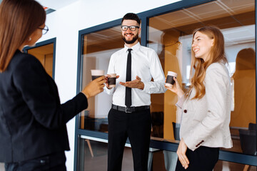 Group of attractive business people standing and communicating together, holding cups, in a modern office. Coffee break.