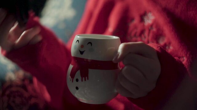 girl in Christmas pajamas drinking coffee in a cup shaped like a snowman. teenager on blue sofa warming his hands with a hot drink. hot milk in cup and hands, woman with tea.
