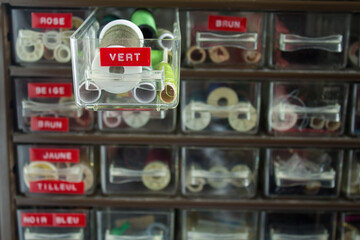 Sewing and tailoring workshop, home sewing, small shelf with colored threads and sewing accessories, the shelf of green threads is open, the names of the colors are in French.