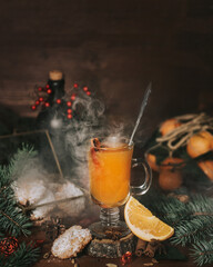 A glass with orange bitter grok stands surrounded by spruce branches and New Year decorations. A box of Christmas cookies. Christmas hot alcoholic drink. Rum bottle. Warming drink. Spices for the grok