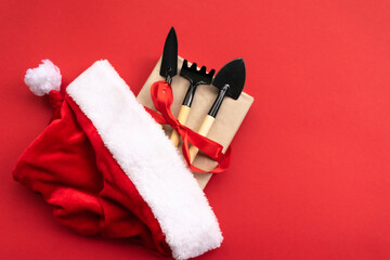 Christmas and new year composition with garden tools, shovel and rake and Santa Claus hat on a red...