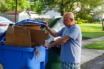Bald senior citizen man standing in a street with his hand on a full trash can