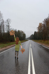 A girl in a green dress with autumn leaves in her hands is walking along the highway