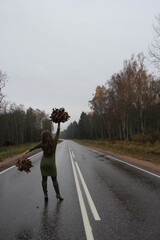 A girl in a green dress with autumn leaves in her hands is walking along the highway