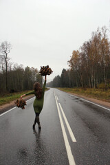A girl in a green dress with autumn leaves in her hands is walking along the highway