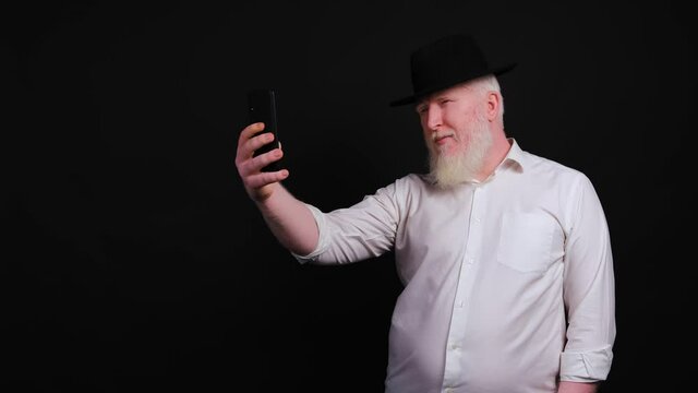 Old man with albinism using phone and taking selfie, making faces on camera, isolated on black background.