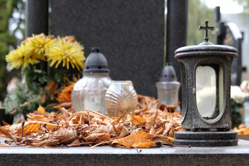 Glass candles and lantern among dry autumn leaves on grave in cemetery on November day