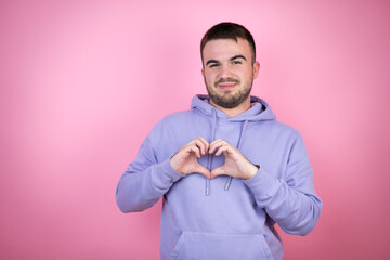 Young handsome man wearing casual sweatshirt over isolated pink background smiling in love showing heart symbol and shape with hands