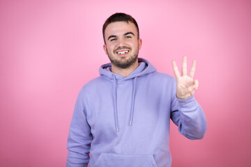 Young handsome man wearing casual sweatshirt over isolated pink background showing and pointing up with fingers number three while smiling confident and happy