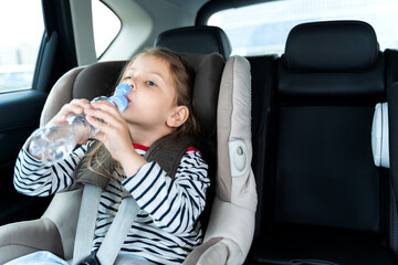 Little caucasian girl is driving in car, dreanking pure water from plastic kids bottle. Traveling on road in safe baby seats with child belts. Fun family trip, activity with parents