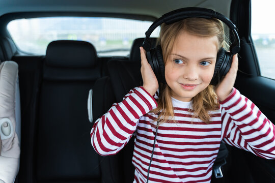 Little Cute Caucasian Girl Is Driving In Car.Kid Listening To Music With Big Headphones.Traveling,riding On Road In Safe Baby Seat With Child Belt.Fun Family Trip,entertainment, Activity With Parents