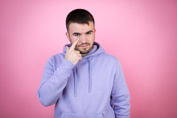 Young handsome man wearing casual sweatshirt over isolated pink background Pointing to the eye watching you gesture, suspicious expression