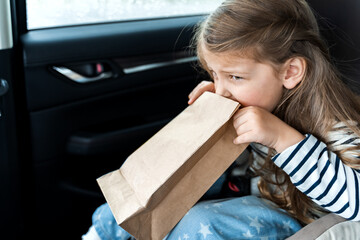 Little girl is driving in car. Kid is sick, feels bad and vomiting into paper bag. Traveling,...