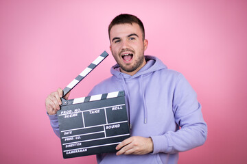 Young handsome man wearing casual sweatshirt over isolated pink background holding clapperboard...