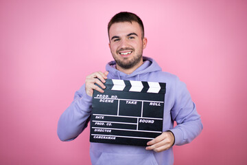 Young handsome man wearing casual sweatshirt over isolated pink background holding clapperboard...