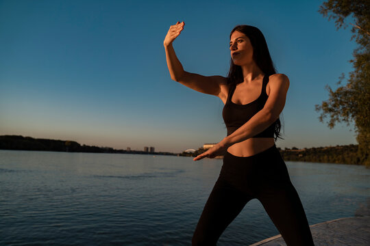 Young adult woman doing tai chi exercises by the river at sunset, wellbeing and health 