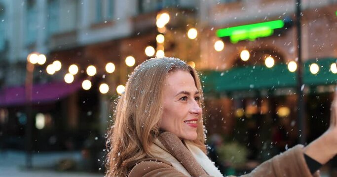 Close up portrait of happy adult Caucasian female in good mood taking selfie photos on smartphone while standing in decorated town and smiling. Joyful Caucasian woman taking pictures on cellphone.