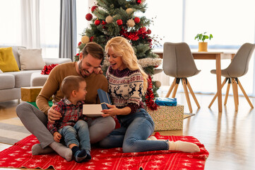 Parents and cute little boy unboxing present in front of Christmas tree. Christmas and holiday season concept