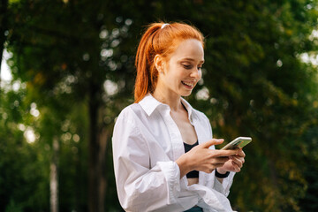 Attractive young red-haired woman wearing sportwear chatting and typing on smartphone with interest...