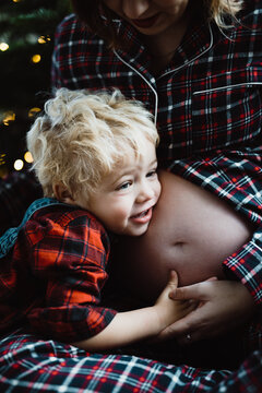 toddler (little boy) hugging the pregnant belly of his mother wearing pyjamas in front of the christmas tree with christmas lights