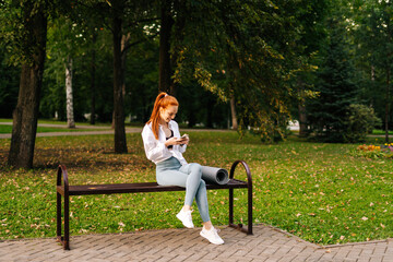 Red-haired joyful young woman wearing sportswear watching video on smartphone sitting bench at city...