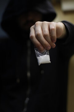 The hand of a hooded drug dealer, as he holds out a transparent sachet in front of him, with powdered cocaine inside to snort.