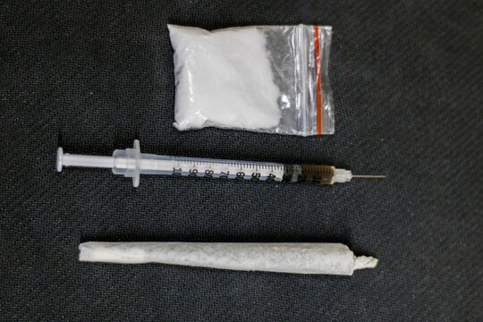 Top view of a syringe full of cocaine, placed in the center between a ganja joint and a cocaine sachet, all placed in contrast to a black surface