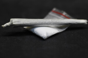 A joint rolled with marijuana or ganja, placed on a sachet full of powdered cocaine, both on a dark...