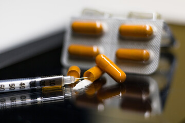 Frontal close-up view of a pack of capsules, one of which is open with a dose of cocaine powder inside, with a syringe positioned next to it, placed on a black and shiny surface.