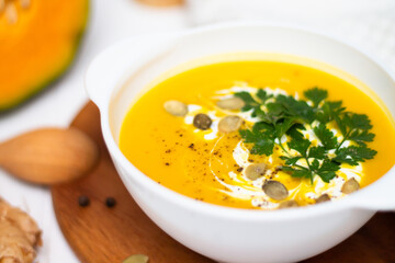 A plate of yellow pumpkin cream soup with cream, herbs and seeds on a white table near raw pumpkin and a wooden spoon.