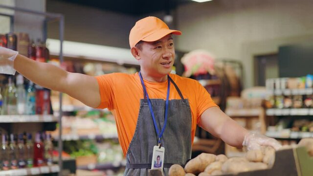 Polite positive asian sellerman worker sorting potatoes in vegetable section helping customer showing right way communicating at supermarket.